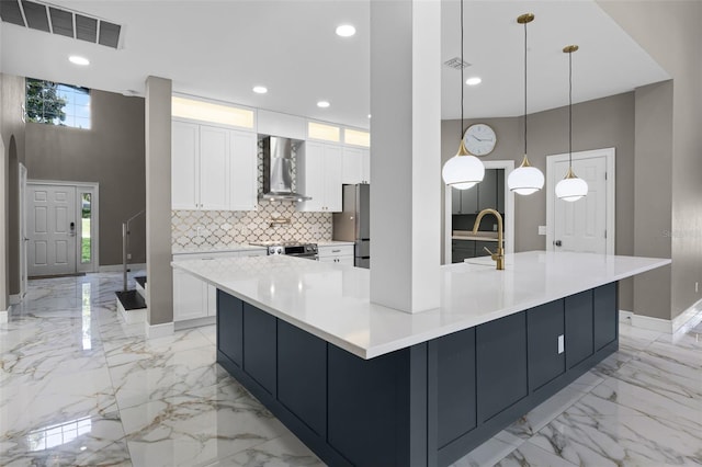 kitchen with wall chimney exhaust hood, light marble finish flooring, appliances with stainless steel finishes, white cabinets, and a towering ceiling