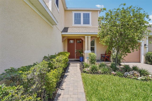 entrance to property with stucco siding, covered porch, driveway, a garage, and roof with shingles