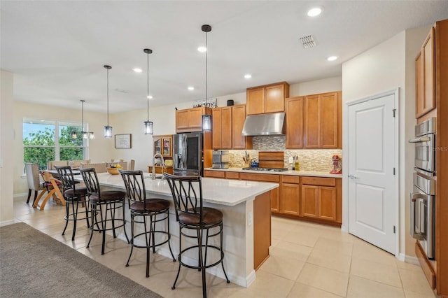 kitchen with under cabinet range hood, stainless steel appliances, backsplash, a center island with sink, and a kitchen bar