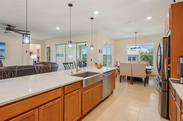 kitchen with a sink, stainless steel appliances, brown cabinetry, a ceiling fan, and light stone counters