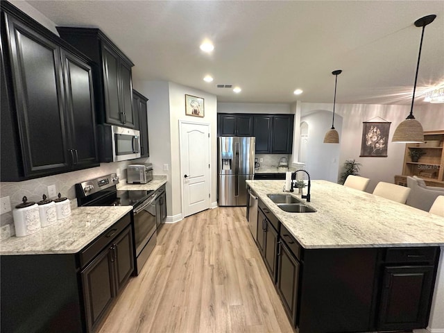 384 Summer Squall Rd, Davenport, FL 33837 - kitchen featuring appliances with stainless steel finishes, dark cabinetry, light wood-style flooring, a center island with sink, and light stone countertops