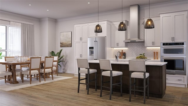 kitchen with stainless steel appliances, wall chimney range hood, hardwood / wood-style floors, an island with sink, and pendant lighting