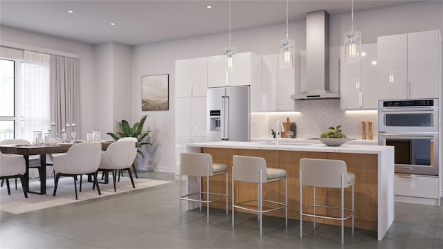 kitchen featuring wall chimney exhaust hood, hanging light fixtures, a kitchen island with sink, white cabinets, and appliances with stainless steel finishes