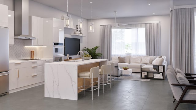 kitchen featuring a kitchen island with sink, wall chimney range hood, sink, decorative light fixtures, and white cabinetry