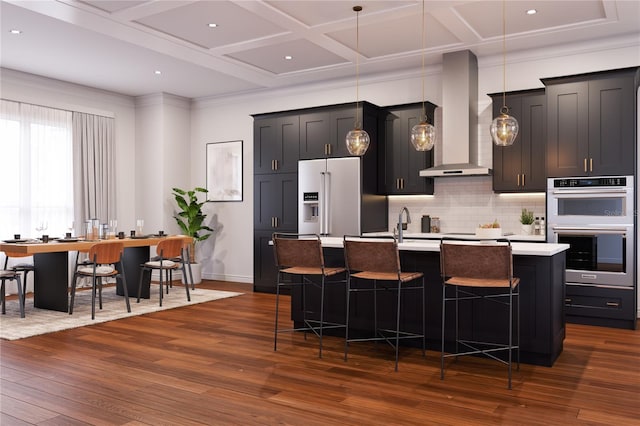 kitchen featuring dark wood-type flooring, wall chimney range hood, an island with sink, a breakfast bar, and appliances with stainless steel finishes