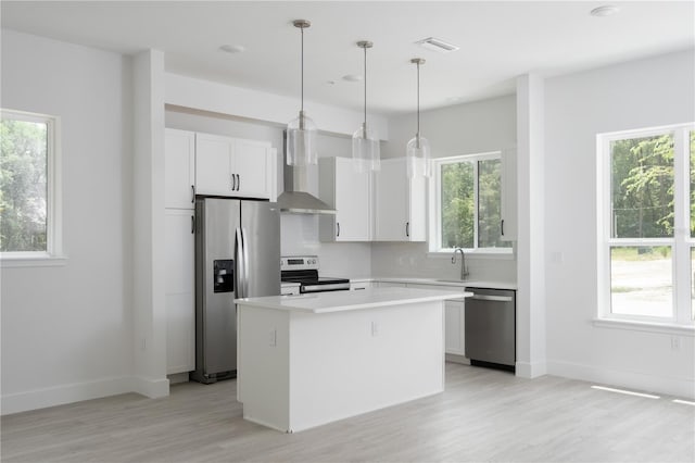 kitchen featuring wall chimney range hood, white cabinetry, stainless steel appliances, a center island, and decorative light fixtures