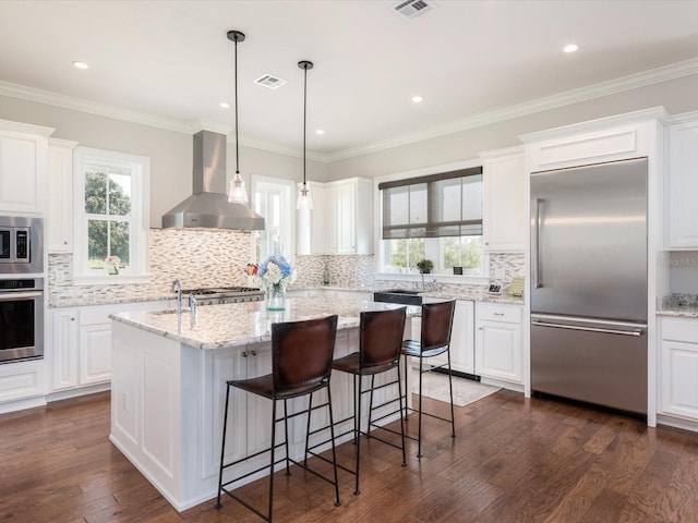 kitchen featuring a kitchen island with sink, white cabinetry, built in appliances, and wall chimney range hood