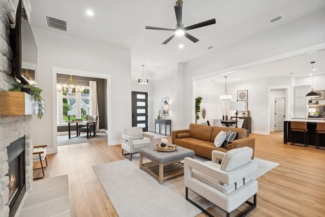 living room with ceiling fan with notable chandelier, light wood-type flooring, and a stone fireplace