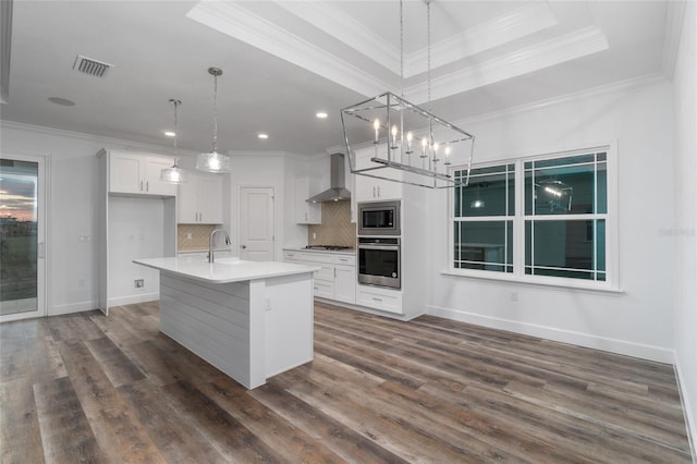 kitchen with an island with sink, a raised ceiling, stainless steel appliances, wall chimney range hood, and decorative light fixtures