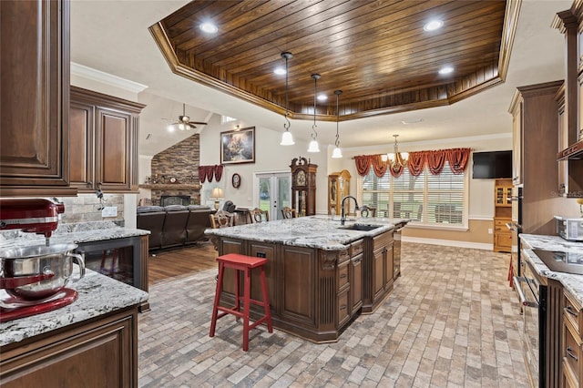 kitchen with an island with sink, sink, a raised ceiling, a fireplace, and ceiling fan with notable chandelier