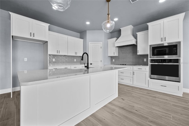 kitchen with appliances with stainless steel finishes, hanging light fixtures, a center island with sink, white cabinetry, and premium range hood