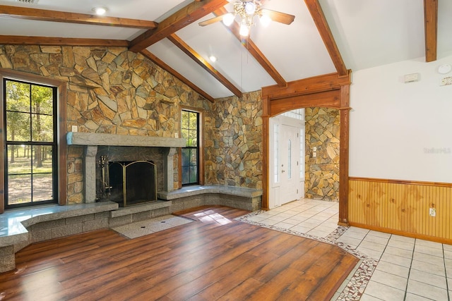 unfurnished living room featuring ceiling fan, light hardwood / wood-style floors, lofted ceiling with beams, and a stone fireplace