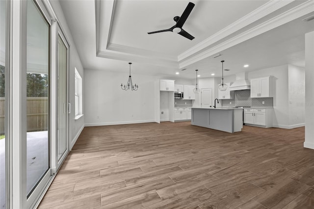 unfurnished living room featuring ceiling fan with notable chandelier, light wood-type flooring, a tray ceiling, ornamental molding, and sink