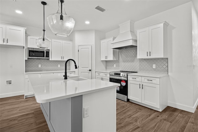 kitchen featuring a kitchen island with sink, white cabinetry, hanging light fixtures, custom range hood, and stainless steel appliances