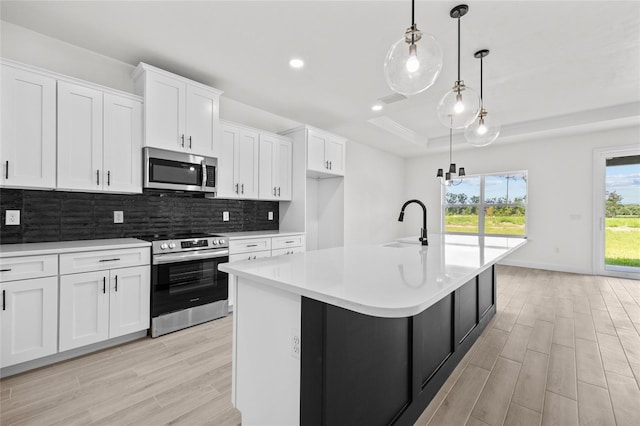 kitchen featuring appliances with stainless steel finishes, a tray ceiling, pendant lighting, white cabinets, and an island with sink