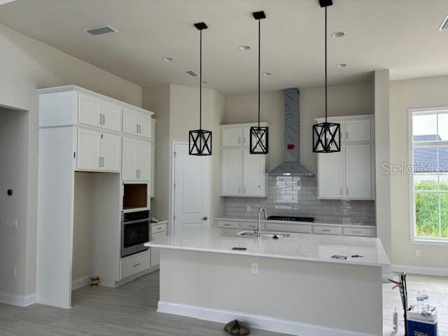 kitchen featuring light wood-type flooring, wall chimney exhaust hood, a kitchen island with sink, hanging light fixtures, and oven