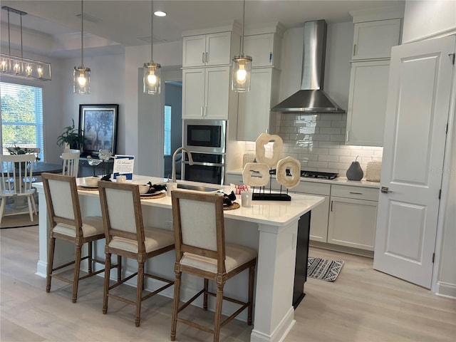 kitchen featuring white cabinetry, wall chimney range hood, light hardwood / wood-style floors, a breakfast bar area, and a center island with sink