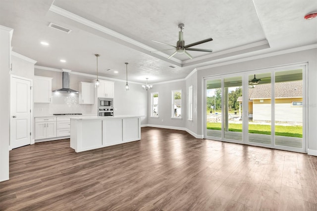 kitchen featuring wall chimney range hood, dark wood-type flooring, stainless steel appliances, an island with sink, and a raised ceiling