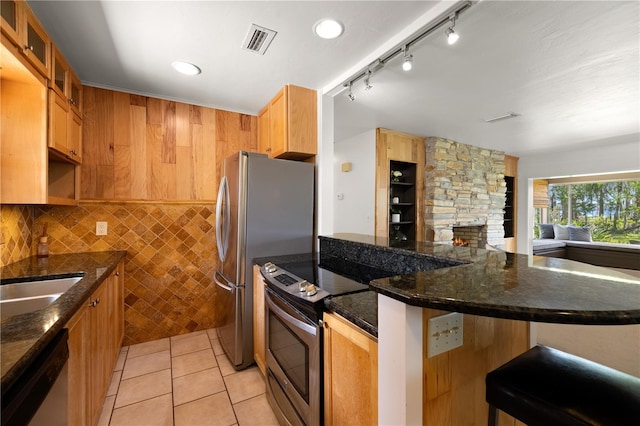 kitchen featuring a fireplace, appliances with stainless steel finishes, dark stone counters, backsplash, and light tile floors