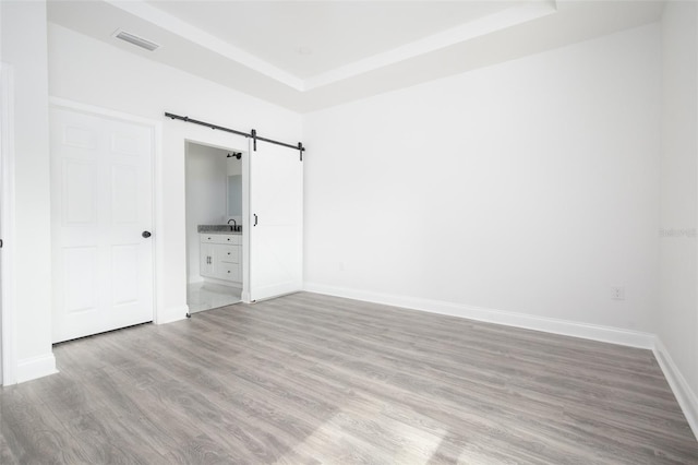 unfurnished bedroom featuring a tray ceiling, light hardwood / wood-style floors, a barn door, and ensuite bathroom