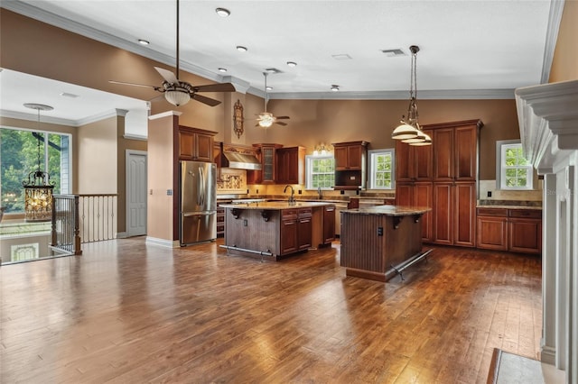 kitchen featuring dark wood-type flooring, a center island, hanging light fixtures, stainless steel refrigerator, and wall chimney range hood