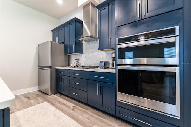 kitchen with decorative backsplash, appliances with stainless steel finishes, wall chimney exhaust hood, blue cabinetry, and light hardwood / wood-style floors