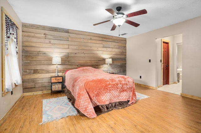 bedroom with ceiling fan, light hardwood / wood-style floors, wooden walls, and ensuite bath