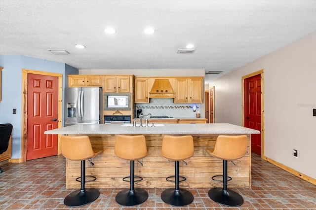 kitchen featuring custom exhaust hood, a center island with sink, appliances with stainless steel finishes, backsplash, and a breakfast bar