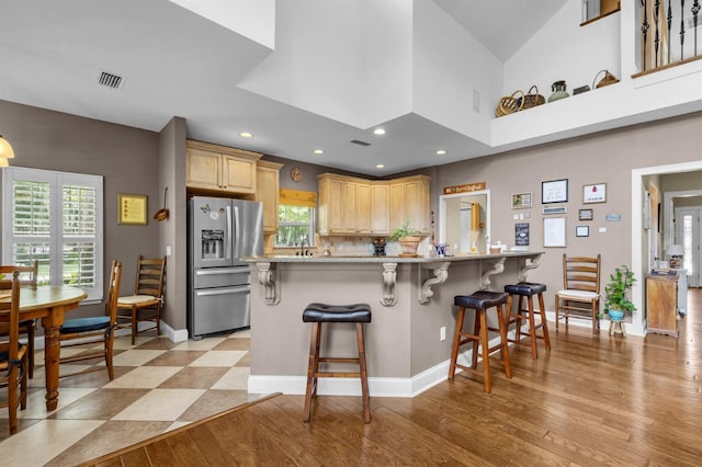 kitchen with a towering ceiling, a kitchen breakfast bar, stainless steel refrigerator with ice dispenser, light brown cabinetry, and light wood-type flooring