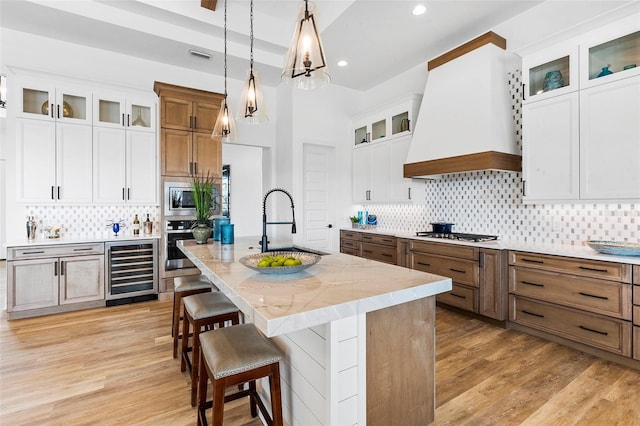 kitchen featuring white cabinetry, custom range hood, light stone countertops, beverage cooler, and a kitchen island with sink