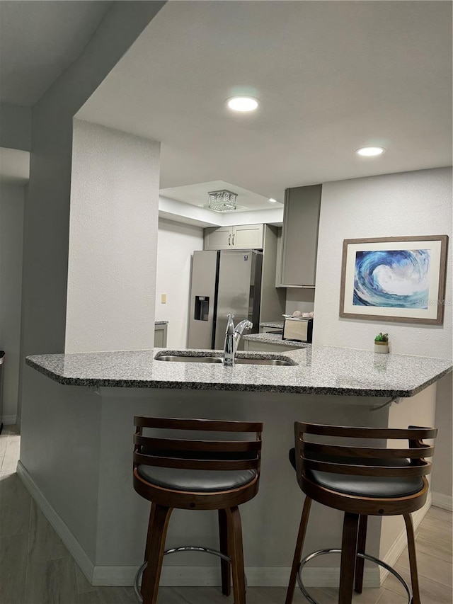 kitchen featuring light stone countertops, gray cabinetry, kitchen peninsula, a breakfast bar area, and stainless steel refrigerator with ice dispenser