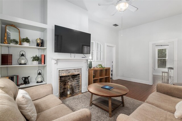 living room with ceiling fan, wood-type flooring, a brick fireplace, and a towering ceiling