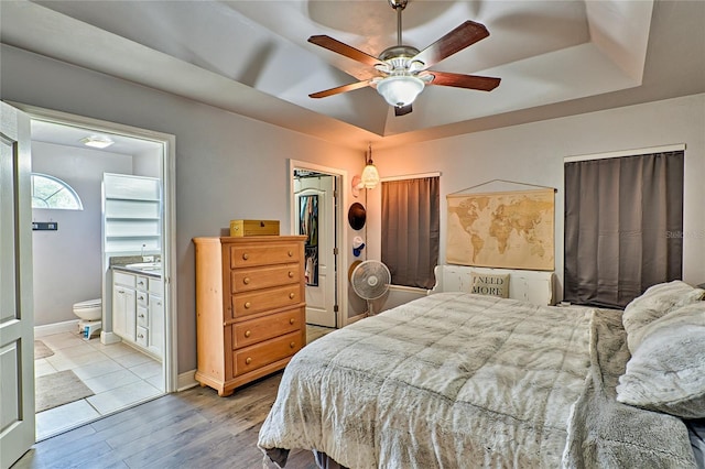 bedroom featuring ensuite bathroom, wood finished floors, a ceiling fan, baseboards, and a tray ceiling