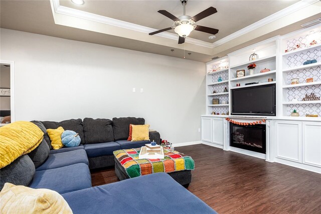 living room with built in shelves, ornamental molding, ceiling fan, a tray ceiling, and dark wood-type flooring