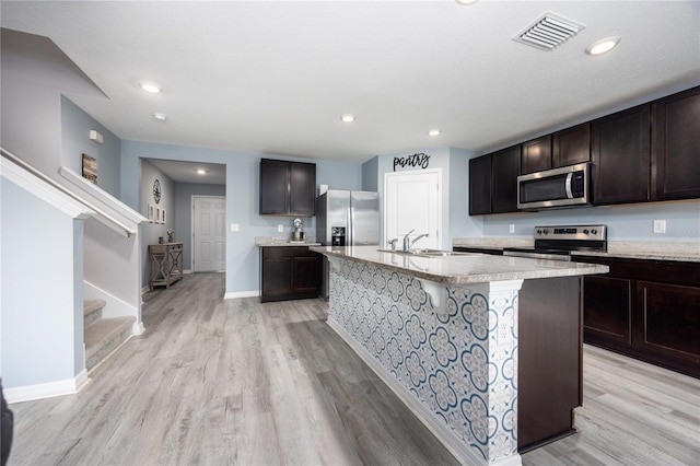 kitchen featuring appliances with stainless steel finishes, an island with sink, light wood-type flooring, dark brown cabinets, and sink