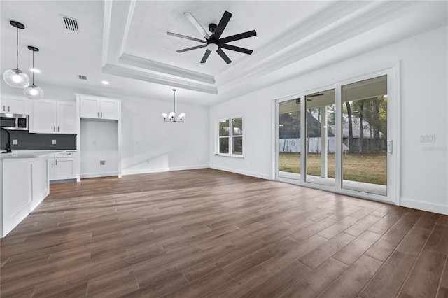 unfurnished living room with a tray ceiling, ceiling fan with notable chandelier, dark wood-type flooring, and a wealth of natural light