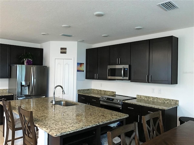 kitchen featuring light stone countertops, stainless steel appliances, a textured ceiling, a kitchen island with sink, and sink