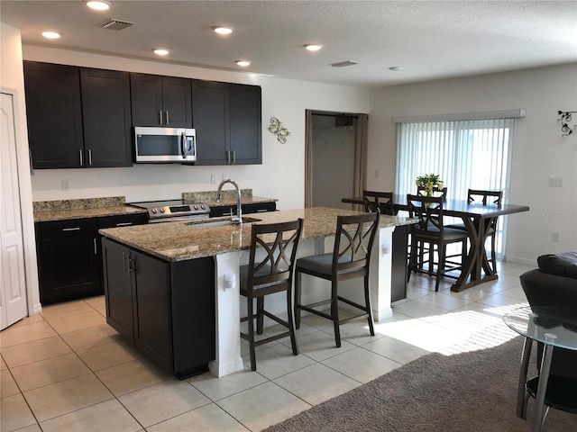 kitchen with light stone countertops, light tile patterned floors, stainless steel appliances, a kitchen island with sink, and sink