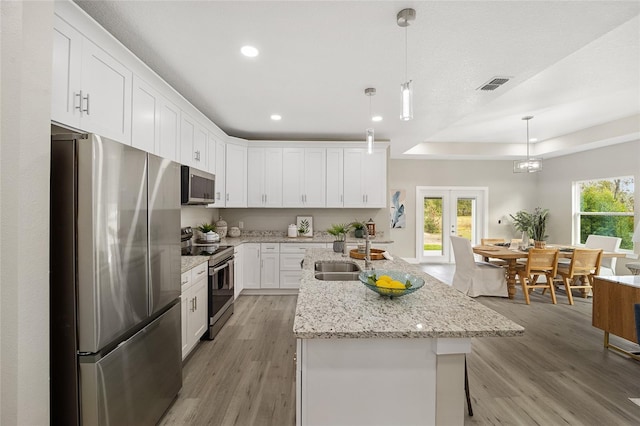 kitchen featuring appliances with stainless steel finishes, light wood-type flooring, a kitchen island with sink, white cabinets, and hanging light fixtures