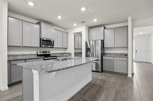 kitchen with appliances with stainless steel finishes, an island with sink, light stone counters, light hardwood / wood-style flooring, and sink