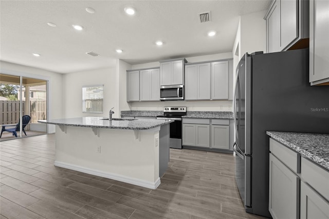 kitchen featuring gray cabinetry, light stone countertops, an island with sink, and stainless steel appliances