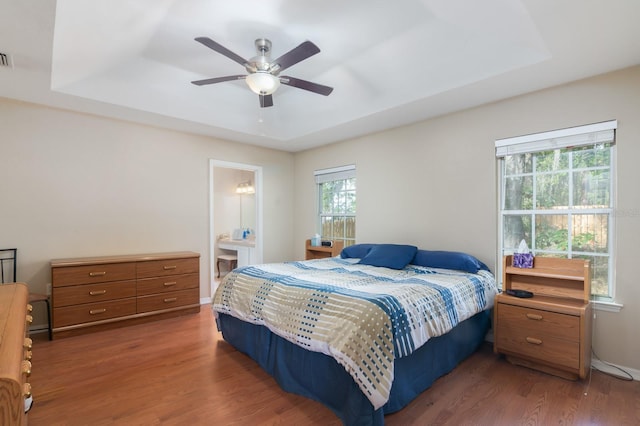 bedroom with dark wood-type flooring, connected bathroom, ceiling fan, and a tray ceiling