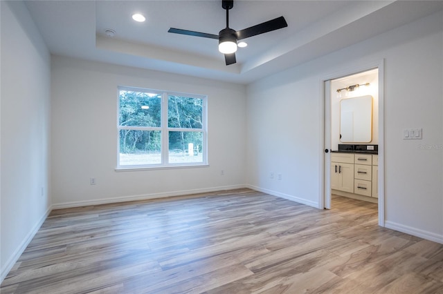 unfurnished bedroom with ensuite bathroom, ceiling fan, a tray ceiling, and light wood-type flooring