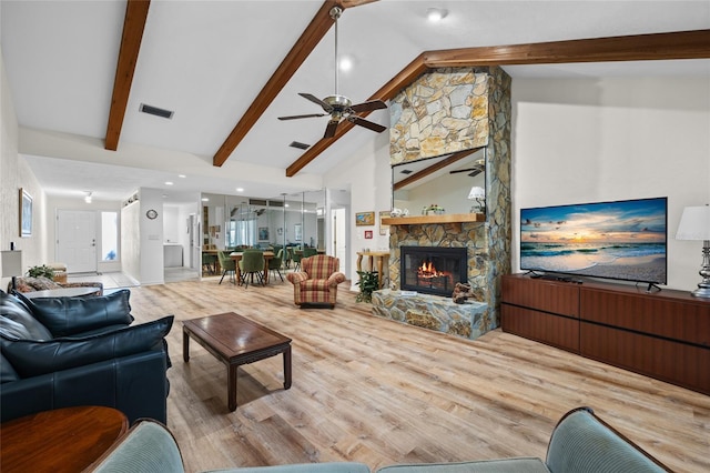 living room with ceiling fan, a stone fireplace, light wood-type flooring, and lofted ceiling with beams