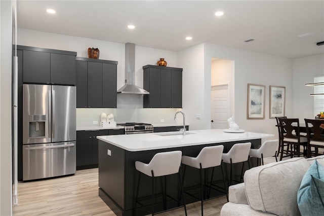 kitchen with sink, a center island with sink, wall chimney range hood, and appliances with stainless steel finishes