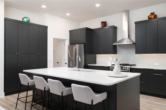 kitchen with sink, wall chimney exhaust hood, light hardwood / wood-style flooring, a kitchen island with sink, and appliances with stainless steel finishes