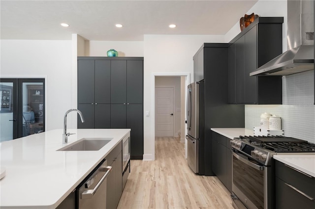 kitchen featuring sink, wall chimney exhaust hood, light hardwood / wood-style flooring, decorative backsplash, and appliances with stainless steel finishes