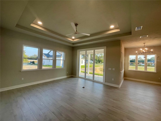 empty room with a raised ceiling, a wealth of natural light, ceiling fan with notable chandelier, and hardwood / wood-style floors