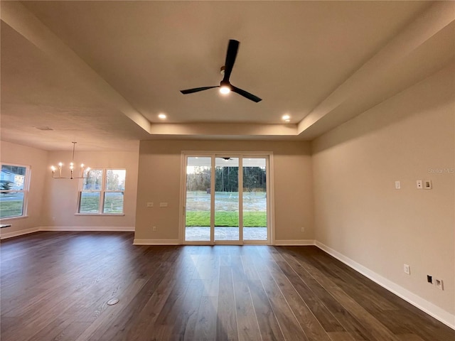 spare room with dark wood-type flooring, a tray ceiling, ceiling fan with notable chandelier, and plenty of natural light