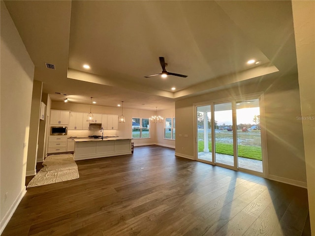 unfurnished living room featuring sink, dark wood-type flooring, a tray ceiling, and ceiling fan with notable chandelier